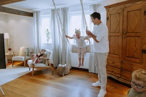 Ein Zimmer mit Holzboden, einem weißen Sofa, einer Schaukel, einem Holzschrank und einem Fenster mit Vorhängen.