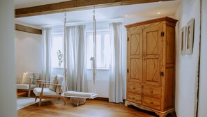 A room with a wooden floor, a white sofa, a swing chair, a wooden cabinet, and a window with curtains. - Studio 28 (Rietberg)