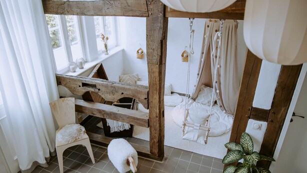 A rustic room with a wooden bed, a white chair, a small table, and a hanging chair. - Studio 28 (Rietberg)