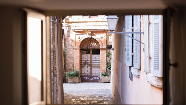 An open door leading to a courtyard with a stone archway, a gate, and potted plants. - Raffaello  (Perugia)