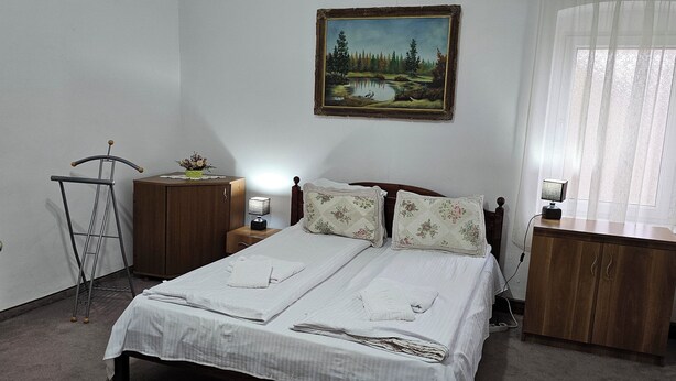Una cama con sábanas blancas impecables, una mesita de noche de madera con una lámpara, un sillón con estampado floral, un armario de madera y un cuadro de un paisaje en la pared.