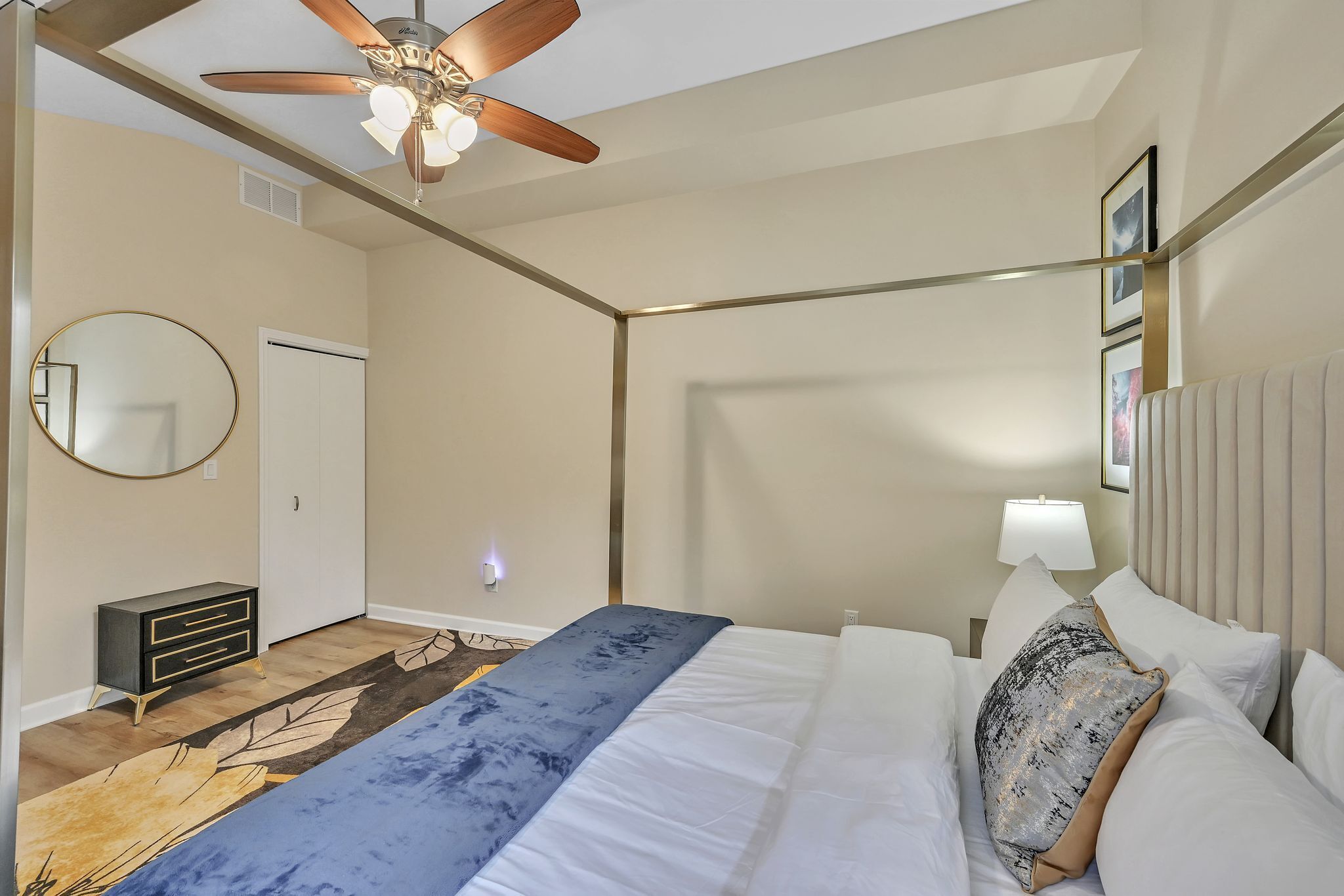 A hotel room with a bed, a ceiling fan, a round mirror, a dresser, and a wall-mounted lamp.