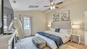 Une chambre à coucher avec une tête de lit capitonnée, un lit avec une literie blanche et bleue, une table de chevet avec une lampe, un ventilateur de plafond et une fenêtre avec des rideaux.