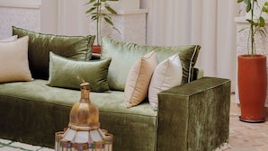 Um sofá verde com várias almofadas, um candeeiro decorativo e um vaso de planta em um quarto com paredes e arcos brancos.