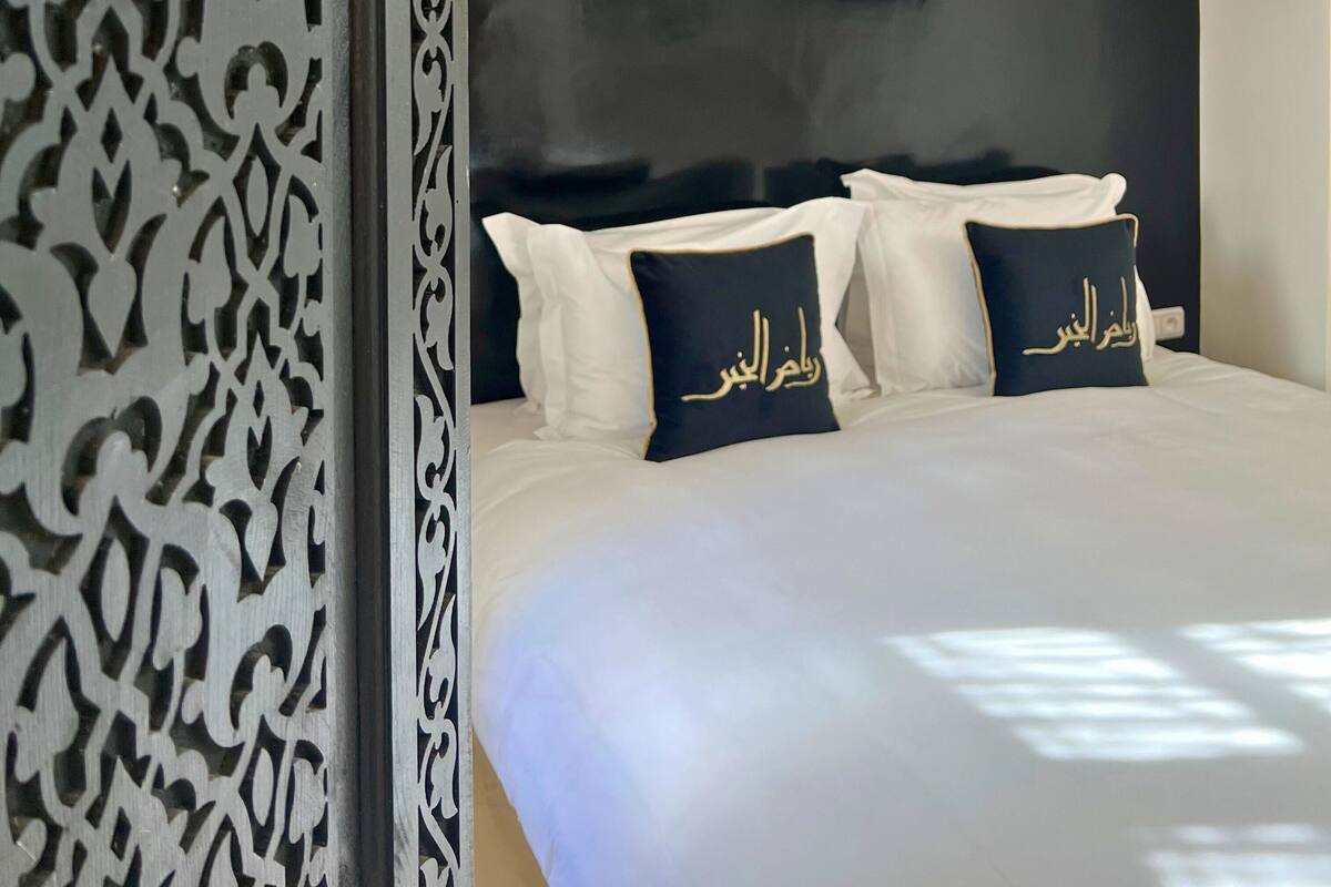 Ein Bett mit schwarzem Kopfteil und weißer Bettwäsche, zwei Kissen mit arabischer Schrift sowie eine Wandverkleidung mit einer Beschriftung.