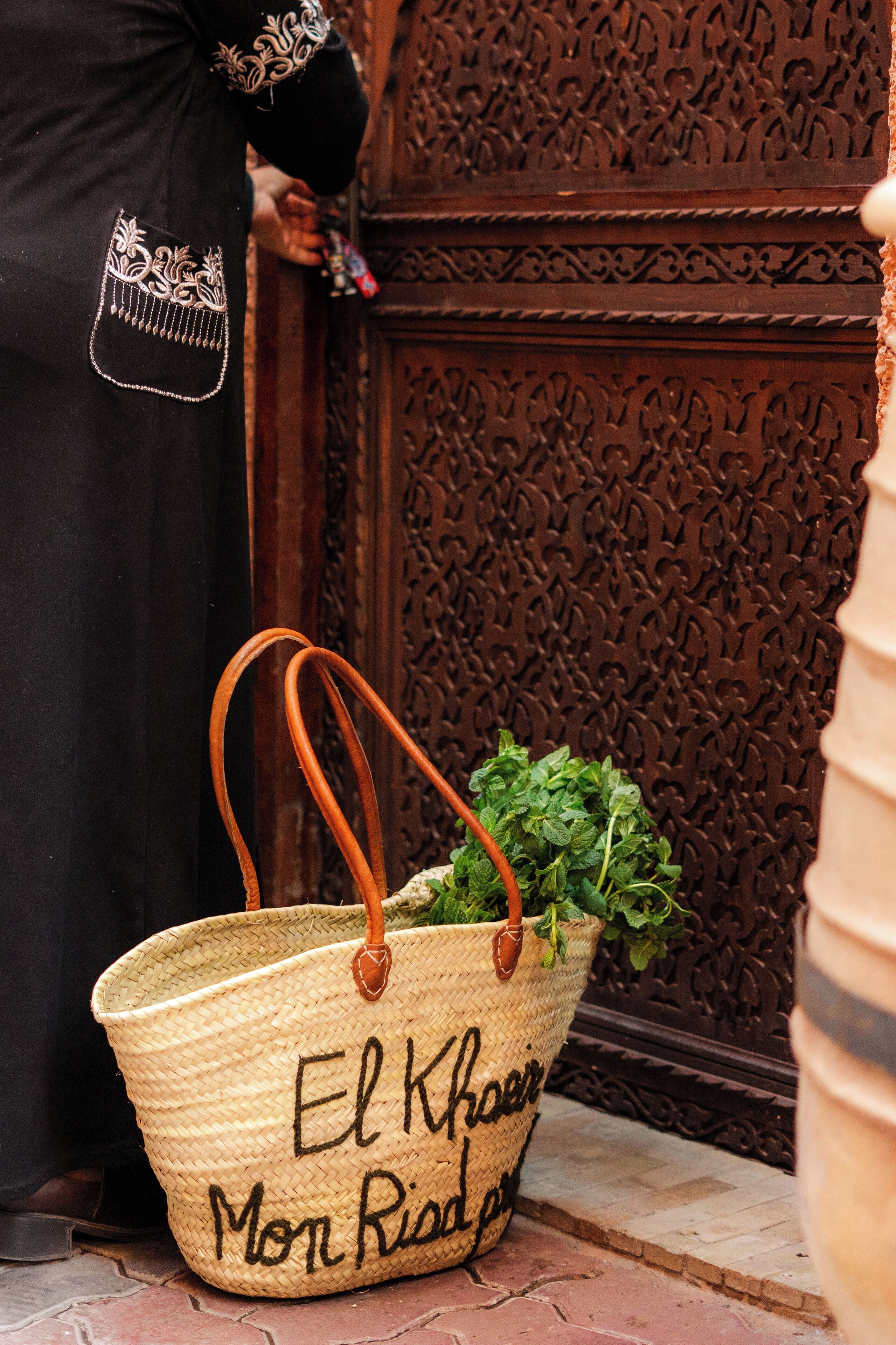 A woven basket with handles and a green plant, placed on a brick-paved surface next to a wooden door with intricate carvings.