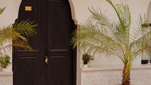 Uma porta de madeira escura com detalhes entalhados, uma grande planta de palmeira em vaso e um candeeiro fixado na parede.