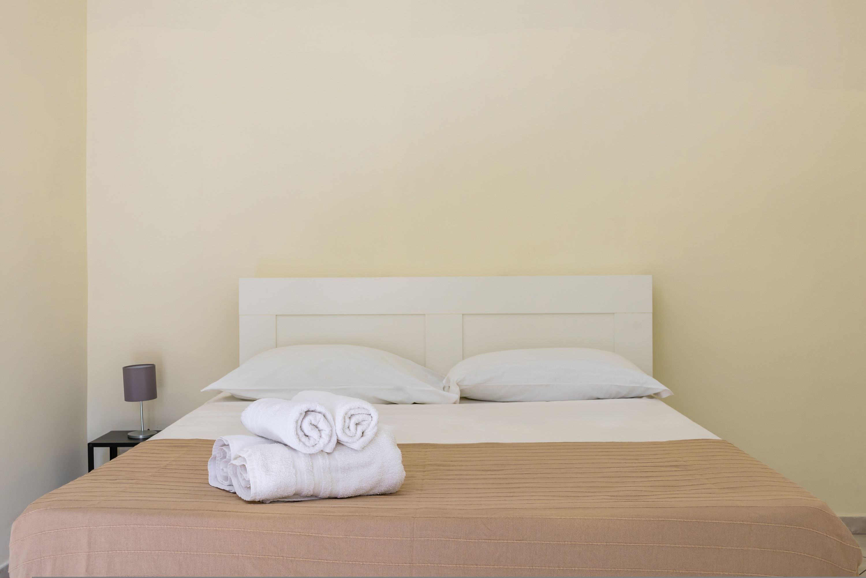Una cama bien hecha con una colcha marrón y dos almohadas blancas, una toalla doblada sobre la cama, y una mesita con una lámpara al lado.