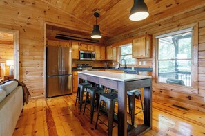 A wooden cabin interior with a kitchen featuring a stainless steel refrigerator, a microwave, and a stove. There is a large island with stools and a window overlooking a deck. - Views @ Whitepath | Mtn Views + Hot Tub + Fire Pit (Ellijay)