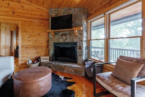 Interior acogedor de una cabaña con chimenea de piedra, paredes de madera, un televisor montado encima y ventanas amplias que ofrecen vistas del exterior.
