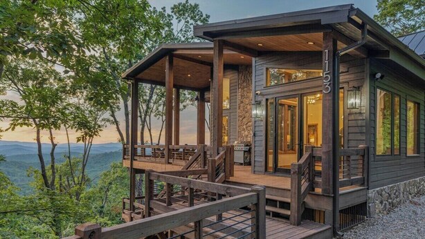 A cabin with a wooden deck and a stone foundation, surrounded by trees and a mountainous backdrop. - Big Abe | Sunset Views + Hot Tub + 6BR Lodge (Blue Ridge)