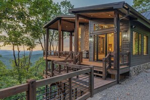 Une cabane dotée d’une terrasse en bois et de fondations en pierre, entourée d’arbres et s’ouvrant sur un paysage montagneux.