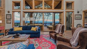 A spacious living area with a large window overlooking a scenic view, furnished with a blue sofa, a brown armchair, and a patterned rug. - Big Abe | Sunset Views + Hot Tub + 6BR Lodge (Blue Ridge)