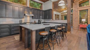 A modern kitchen with dark cabinetry, a large island with stools, and large windows overlooking a forested area. - Big Abe | Sunset Views + Hot Tub + 6BR Lodge (Blue Ridge)