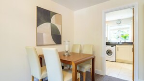 A dining area with a table and chairs, a wall-mounted abstract painting, and a view into a kitchen with a washing machine. - Shrewsbury Apartment (Shrewsbury)