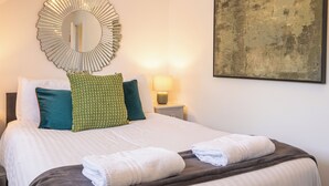 A neatly made bed with pillows, a round mirror, a bedside lamp, and a framed artwork on the wall. - Shrewsbury Apartment (Shrewsbury)