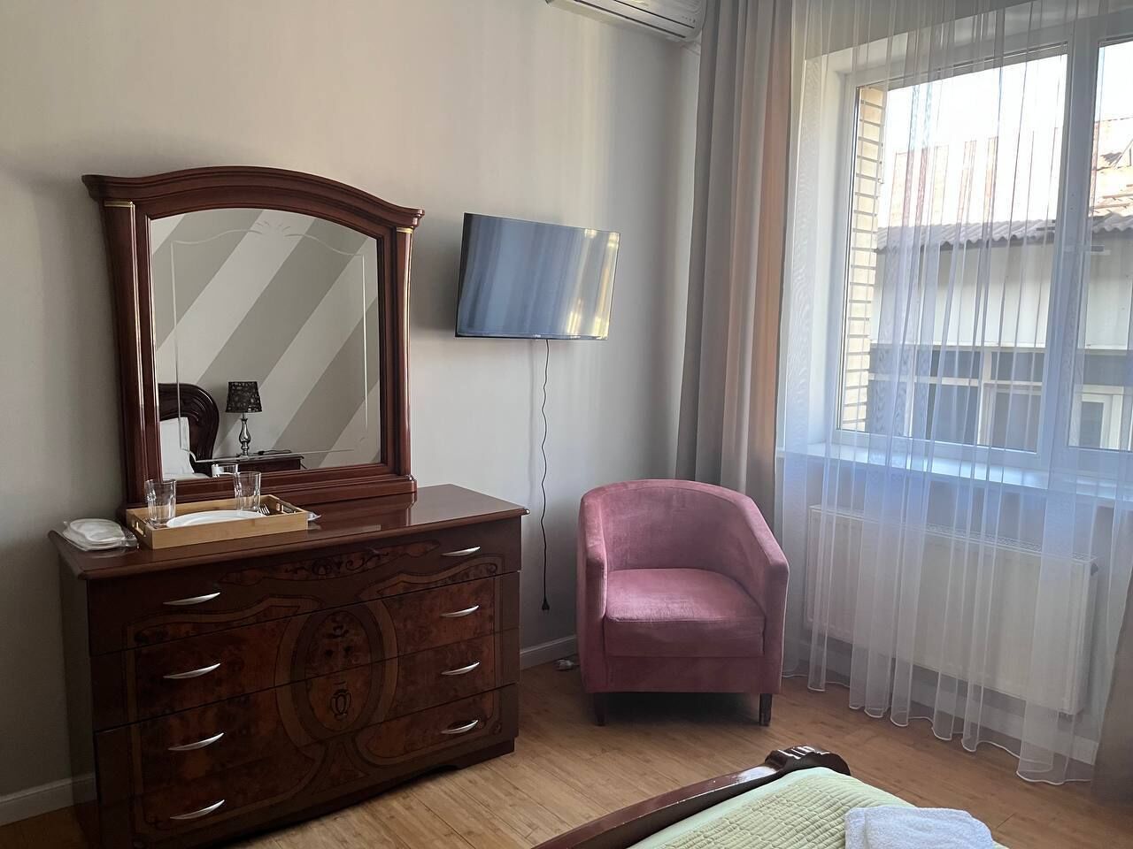 A bedroom with a wooden dresser, a pink chair, a flat-screen TV, and a window with sheer curtains.