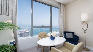 Een moderne hotelkamer met een groot raam met uitzicht op de stad, een ronde witte tafel, twee beige fauteuils, een blauwe vaas met bloemen en een vloerlamp.