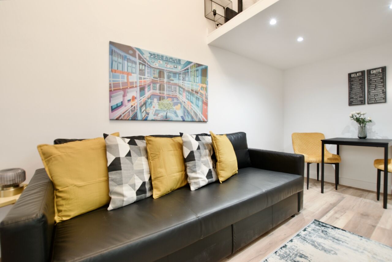 Ein modernes Wohnzimmer mit einem schwarzen Ledersofa, senfgelben und gemusterten Kissen, einem Stadtbildgemälde, einer kleinen Essecke mit Tisch und Stühlen und einer Kaffeekarte an der Wand.