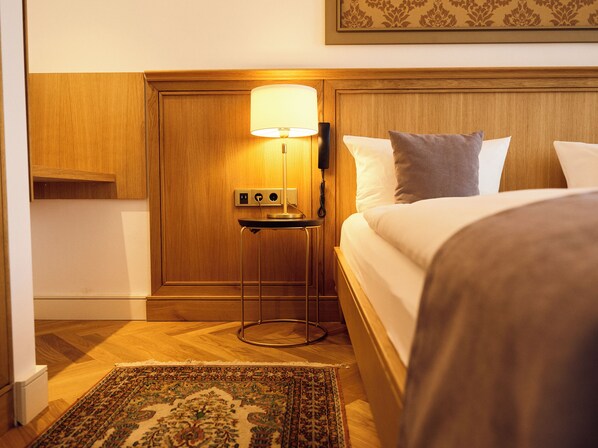 Habitación de hotel con cama, mesita de noche con lámpara, toma de corriente y alfombra estampada.