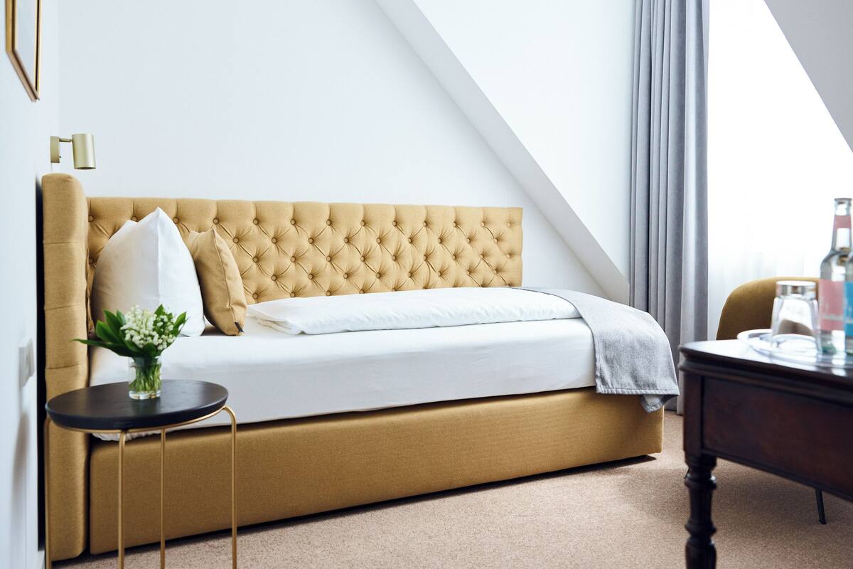 A modern bedroom with a tufted headboard sofa, a small round table with a vase, and a view of the sky through a window.