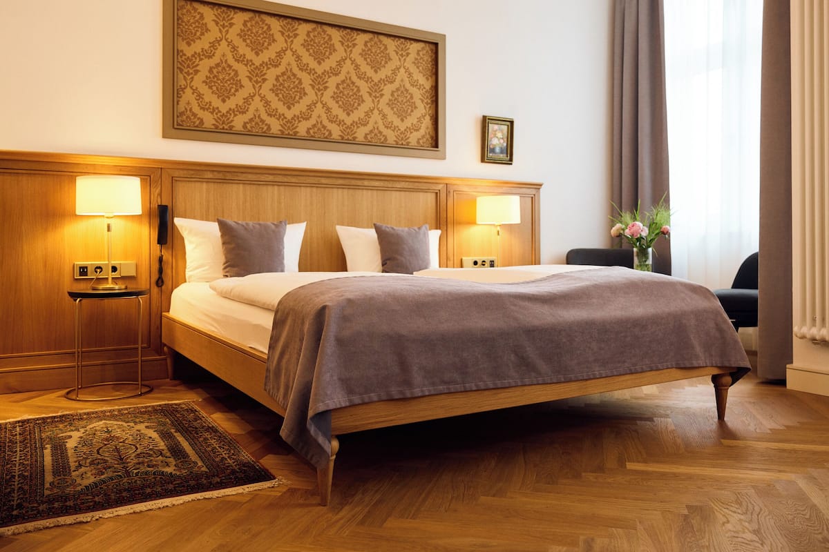 A hotel room with a large bed, two bedside lamps, a wooden headboard, a patterned wall hanging, a small table with a lamp, a vase with flowers, and a view of a window with curtains.