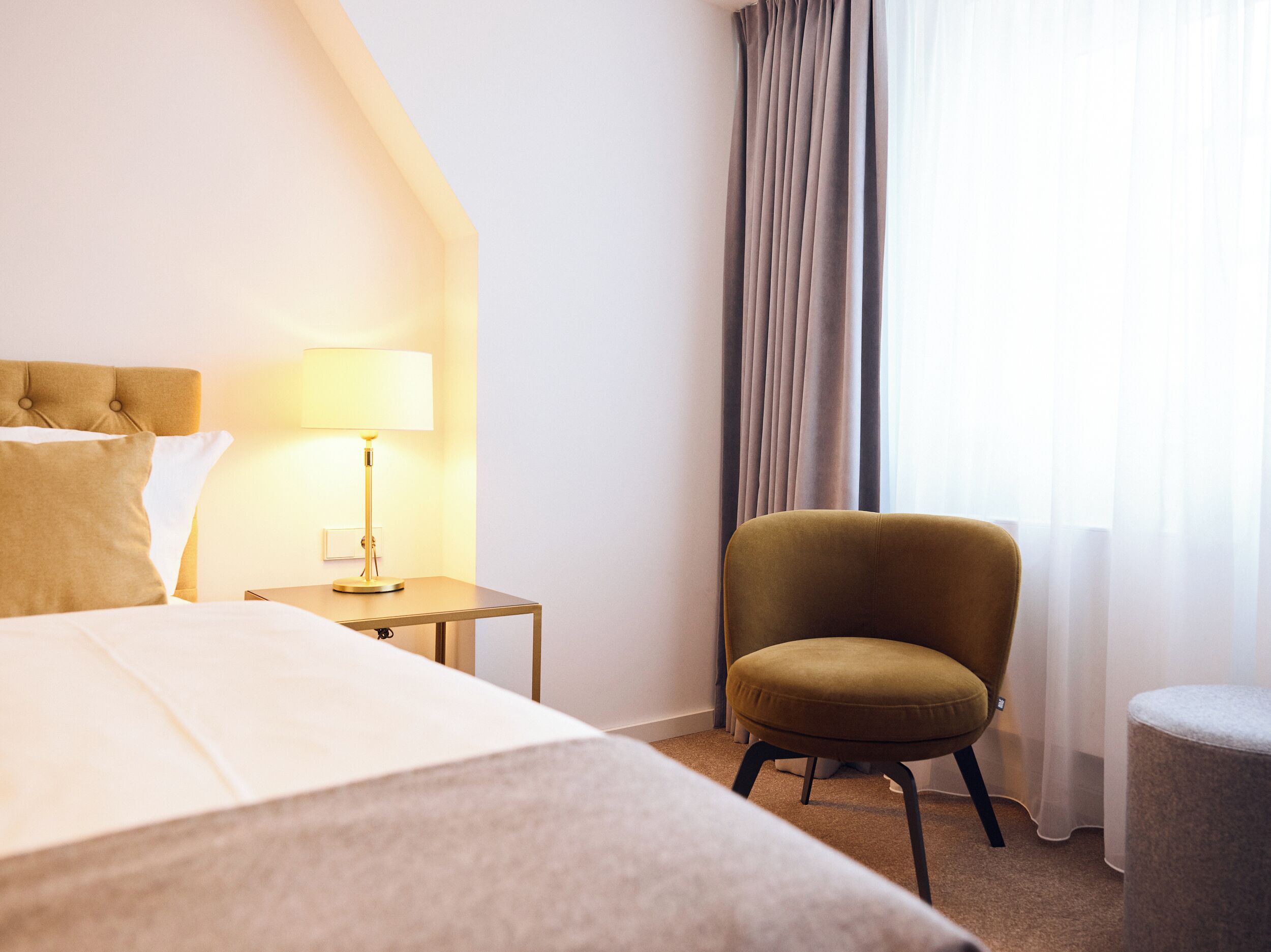 A hotel room with a bed, a side table with a lamp, a chair, and a window with curtains.