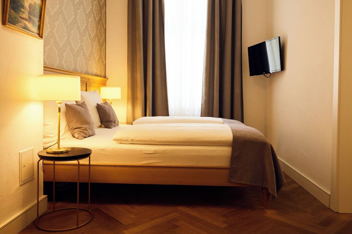 A hotel room with a bed, bedside lamps, a television on the wall, and a painting on the wall.