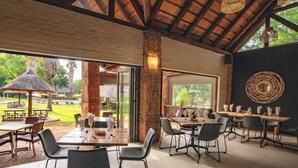 An outdoor dining area with tables and chairs, a thatched-roof structure, and a view of a pool and greenery. - Olifants River Camping by Dream Resorts (Middelburg)