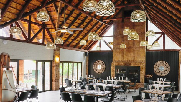A spacious dining area with high ceilings, exposed wooden beams, and multiple hanging light fixtures. Tables are set with white tablecloths and black chairs. - Olifants River Camping by Dream Resorts (Middelburg)