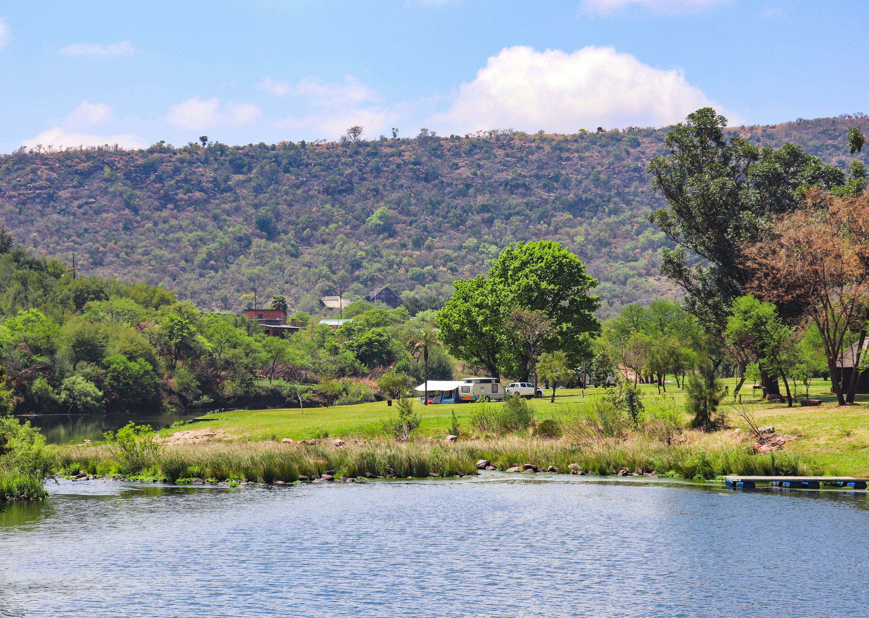 Vista a la orilla del río con una casa, un árbol y una colina al fondo.