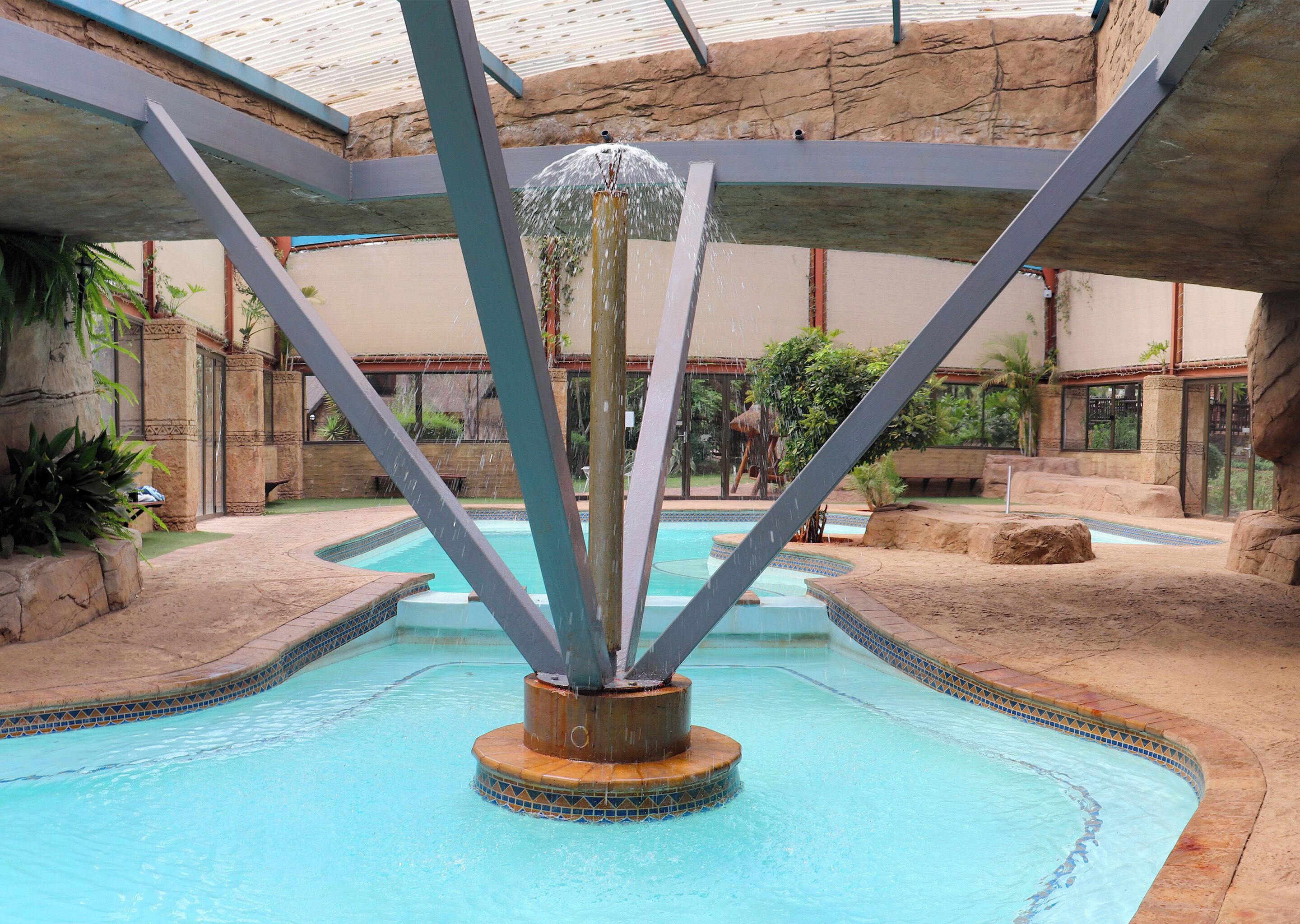 An indoor pool with a central fountain, surrounded by stone walls and tropical plants.
