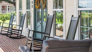 En overdækket veranda med røde mursten, sorte metalstole, en ventilator i loftet og en dør pyntet med en krans.