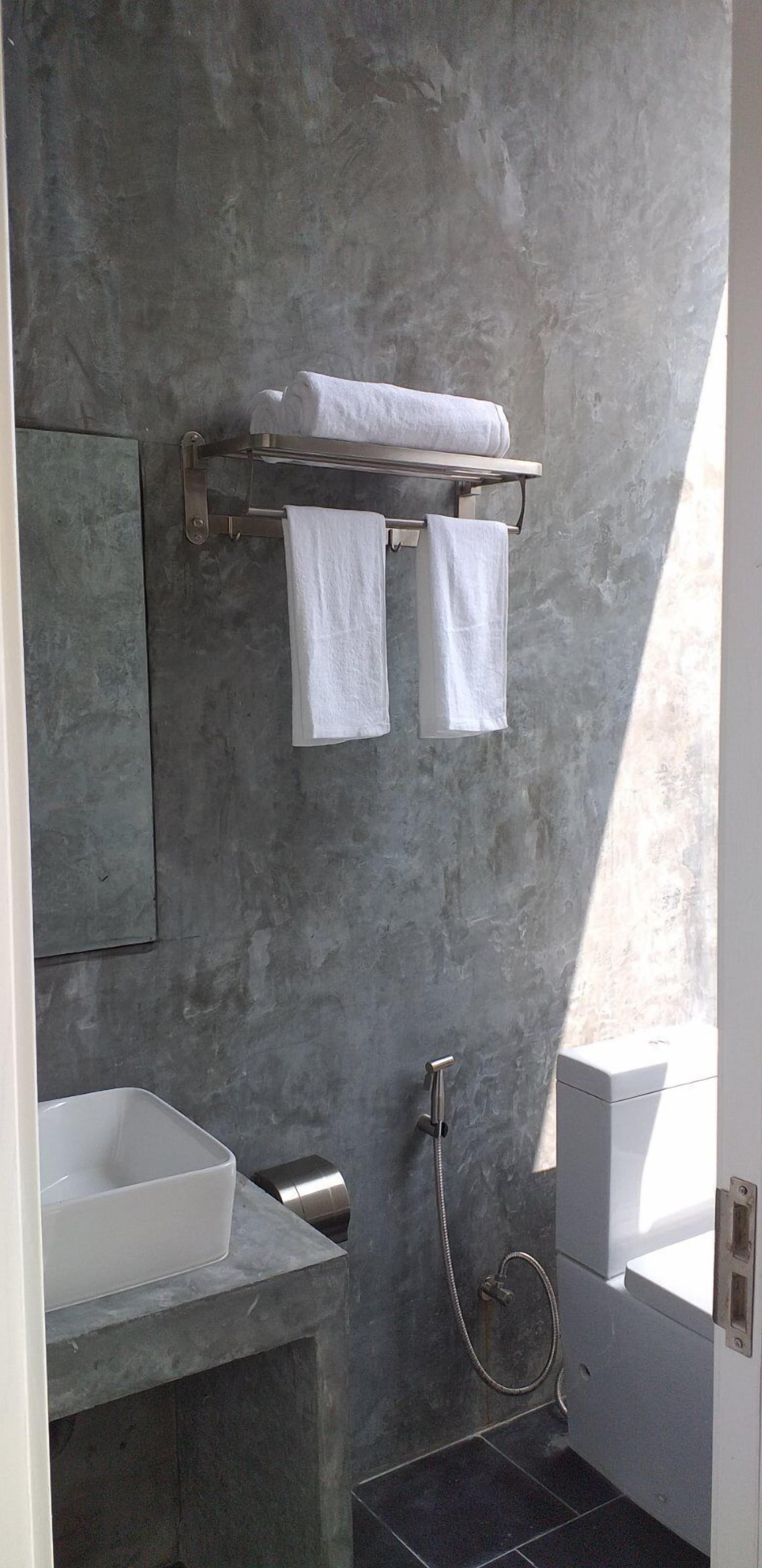 Un baño moderno con una pared de hormigón gris, un lavabo blanco, un perchero con dos toallas, un cabezal de ducha de mano y un inodoro blanco.