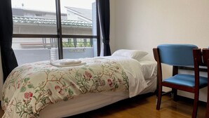 A neatly made bed with a floral bedspread, a wooden chair, and a view of buildings through a window. - W311 Tokyo Monthly rental- Sakurashinmachi Sunrise (Setagaya City)