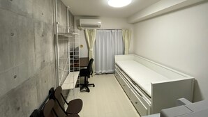 Ein schlichter Raum mit einem Einzelbett, einem Schreibtisch mit Stuhl, einer Schrankwand und einem Vorhang.