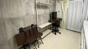 Ein kleiner Raum mit einer Betonwand, einem Schreibtisch mit Stuhl, einem Fernseher und einem Regalsystem.