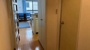 A narrow hallway with wooden flooring leading to a room with a bed, a desk, and a window with blue curtains. - W306 Monthly Rental Tokyo - Sunrise Living (Setagaya City)