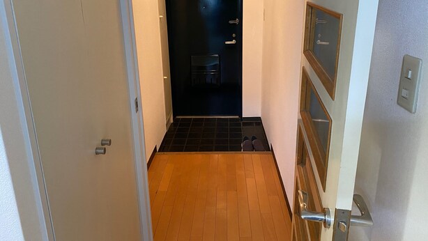 A hallway with a wooden floor, a black door, and a white wall with a light switch. - W306 Monthly Rental Tokyo - Sunrise Living (Setagaya City)