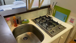 A compact kitchen with a stainless steel sink, a gas stove, and a small wooden stool. - W306 Monthly Rental Tokyo - Sunrise Living (Setagaya City)