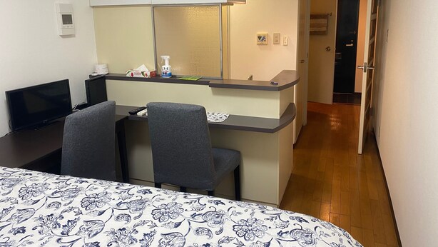 Une chambre d’hôtel compacte comprenant un lit, un bureau avec un ordinateur et une chaise, une petite kitchenette et une salle de bain visible à travers une porte ouverte.