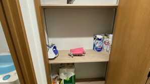 A small storage closet with shelves containing various items including a spare blanket, a pink hairdryer, and several bottles of personal care products. - W212 Tokyo Monthly Rental - Komazawa Telework (Setagaya City)