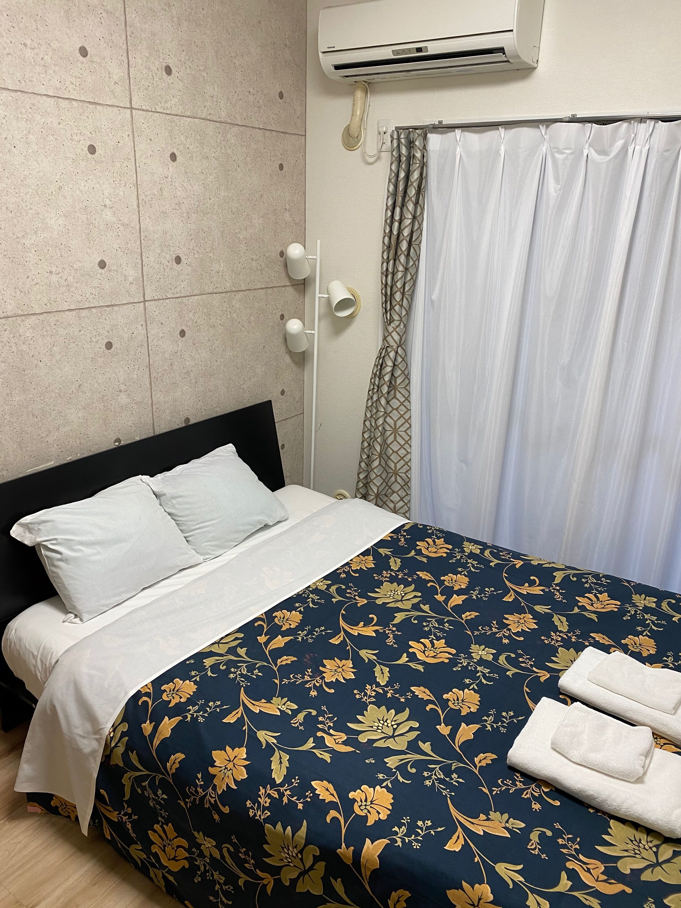 A hotel room with a bed, white curtains, an air conditioning unit, and a wall-mounted light fixture.