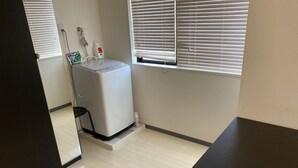 A laundry room with a washing machine, a small trash bin, and a window with blinds. - W201 Monthly Rental Tokyo - Large Studio (Setagaya City)