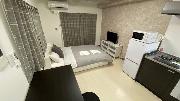 A compact hotel room with a bed, a small desk, a refrigerator, a microwave, and a TV. - W201 Monthly Rental Tokyo - Large Studio (Setagaya City)