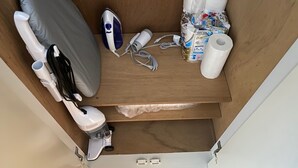Een kleine berging met een matras, een strijkijzer, een stofzuiger en diverse toiletartikelen.