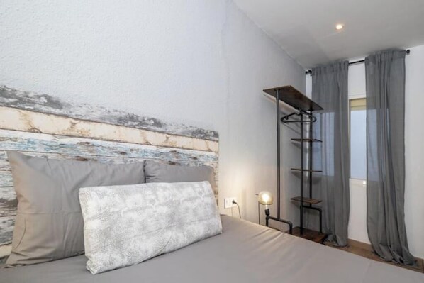 Un dormitorio con cama, almohadas, una mesita de noche, una lámpara y una ventana con cortinas.