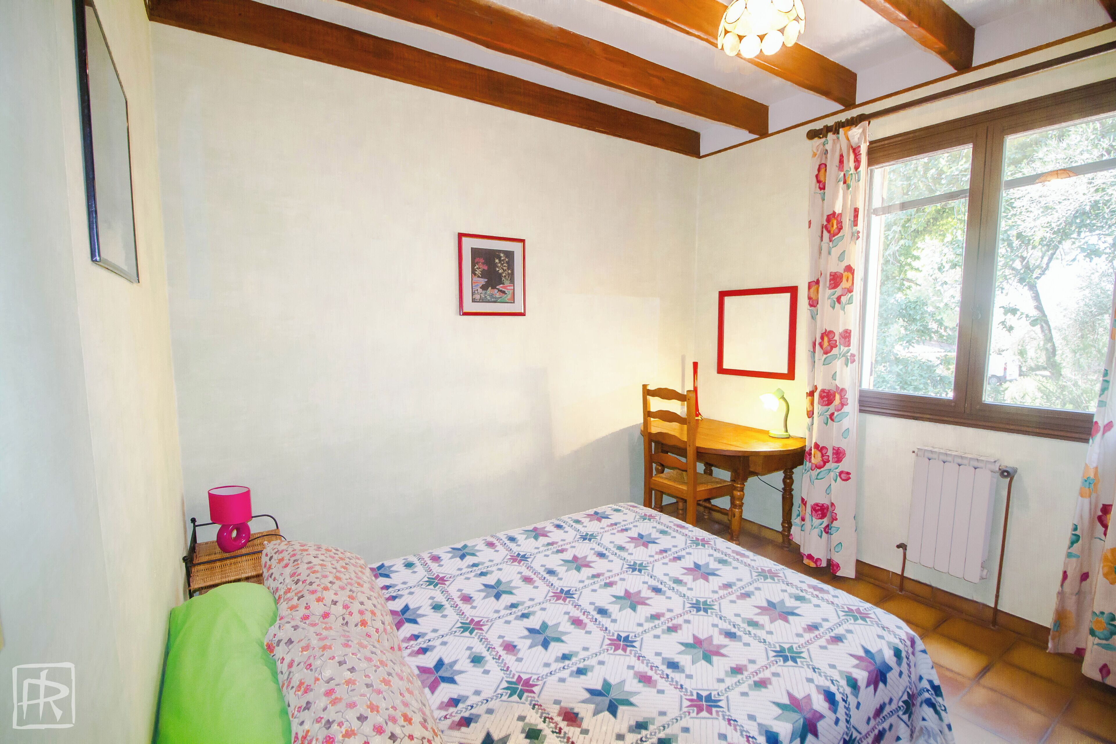 Une chambre à coucher avec un lit, une chaise, une table, une lampe et une fenêtre avec des rideaux.