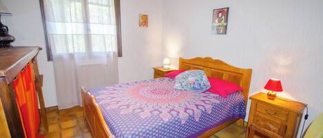 A bedroom with a wooden bed, a nightstand with a lamp, a chair, a window with curtains, and a small table.