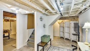 Una habitación amplia con techo alto, vigas a la vista, una escalera, una cesta verde sobre un banco y un escritorio con una lámpara.